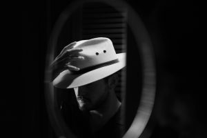A moody black and white portrait of a man adjusting his hat in front of a mirror.
